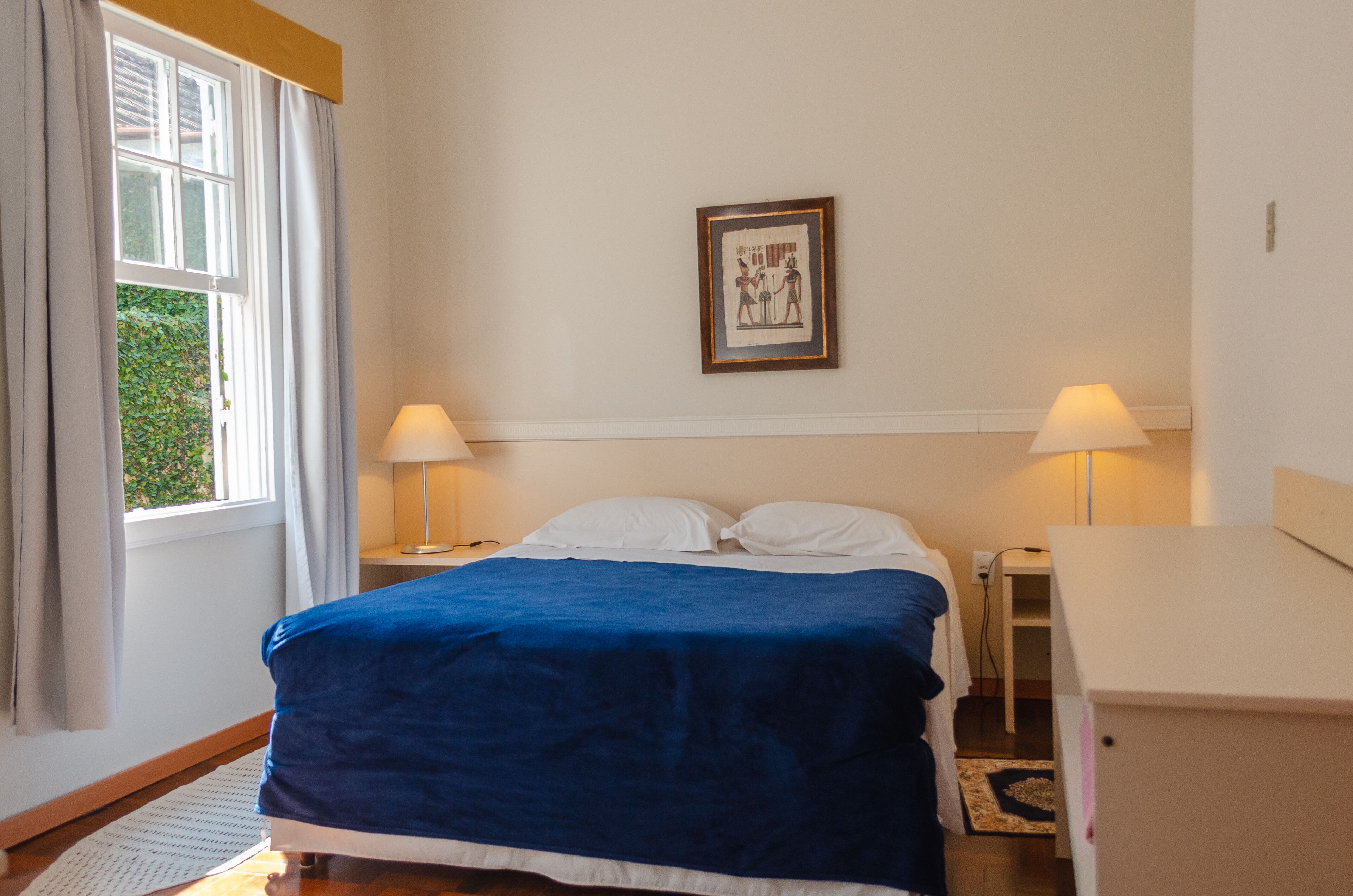 A bedroom with a bed, two bedside lamps, a framed picture on the wall, and a window with curtains.