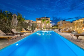 A swimming pool with lounge chairs and umbrellas, surrounded by a fence and a building in the background. - L&C Apartments (Rhodes)