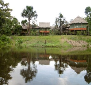 Un entorno tranquilo junto al lago con construcciones de techo de paja, probablemente alojamientos, rodeado de exuberante vegetación.