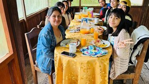Un grupo de personas sentadas alrededor de una mesa con comida y bebidas, en una habitación con paredes de madera y ventanas amplias.