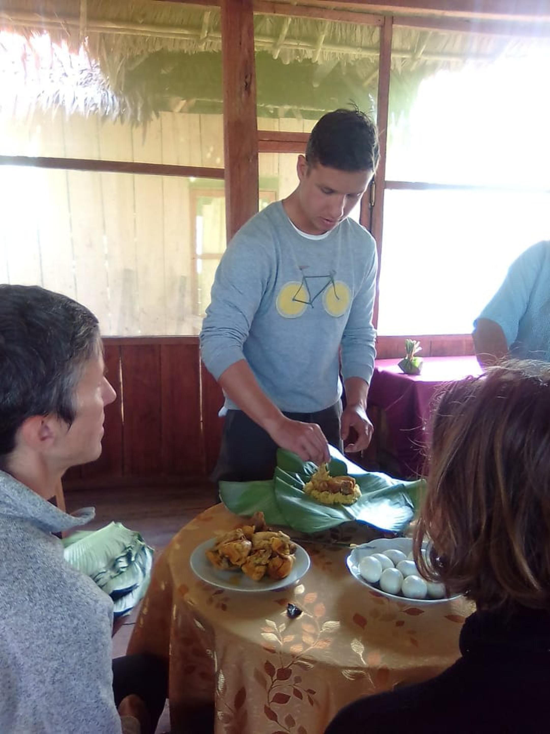 A person serving food at a table with a thatched roof and wooden walls.