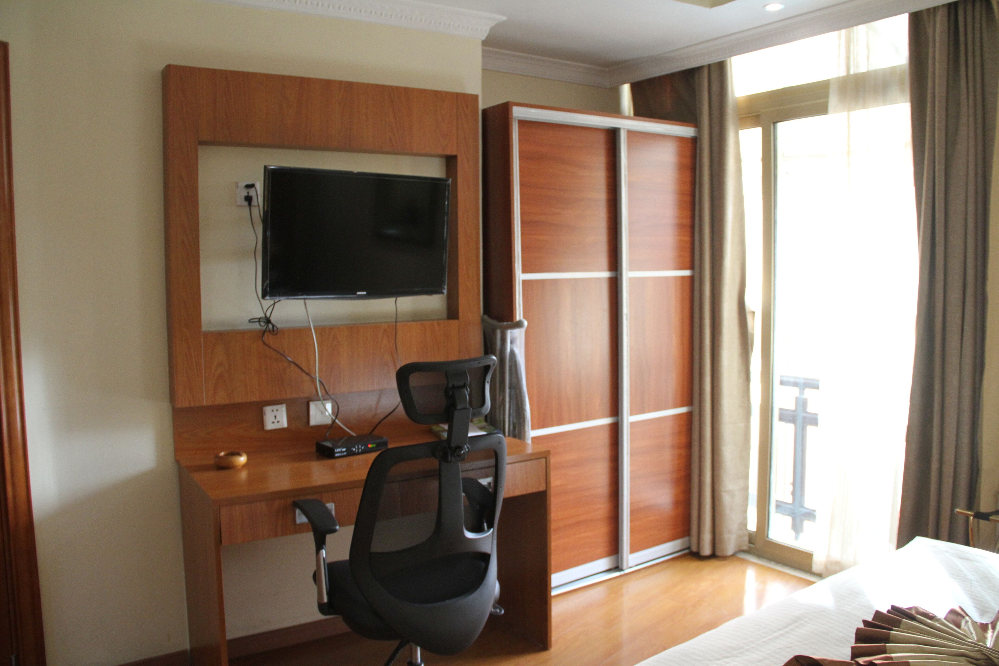 Een hotelkamer met een houten bureau, een televisie erboven, een schuifdeurkast en een groot raam met gordijnen.