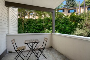 A covered balcony with a table and two chairs, overlooking a garden and buildings. - Seattle Vacation Home: Thomas Lodge Vermont (Seattle)
