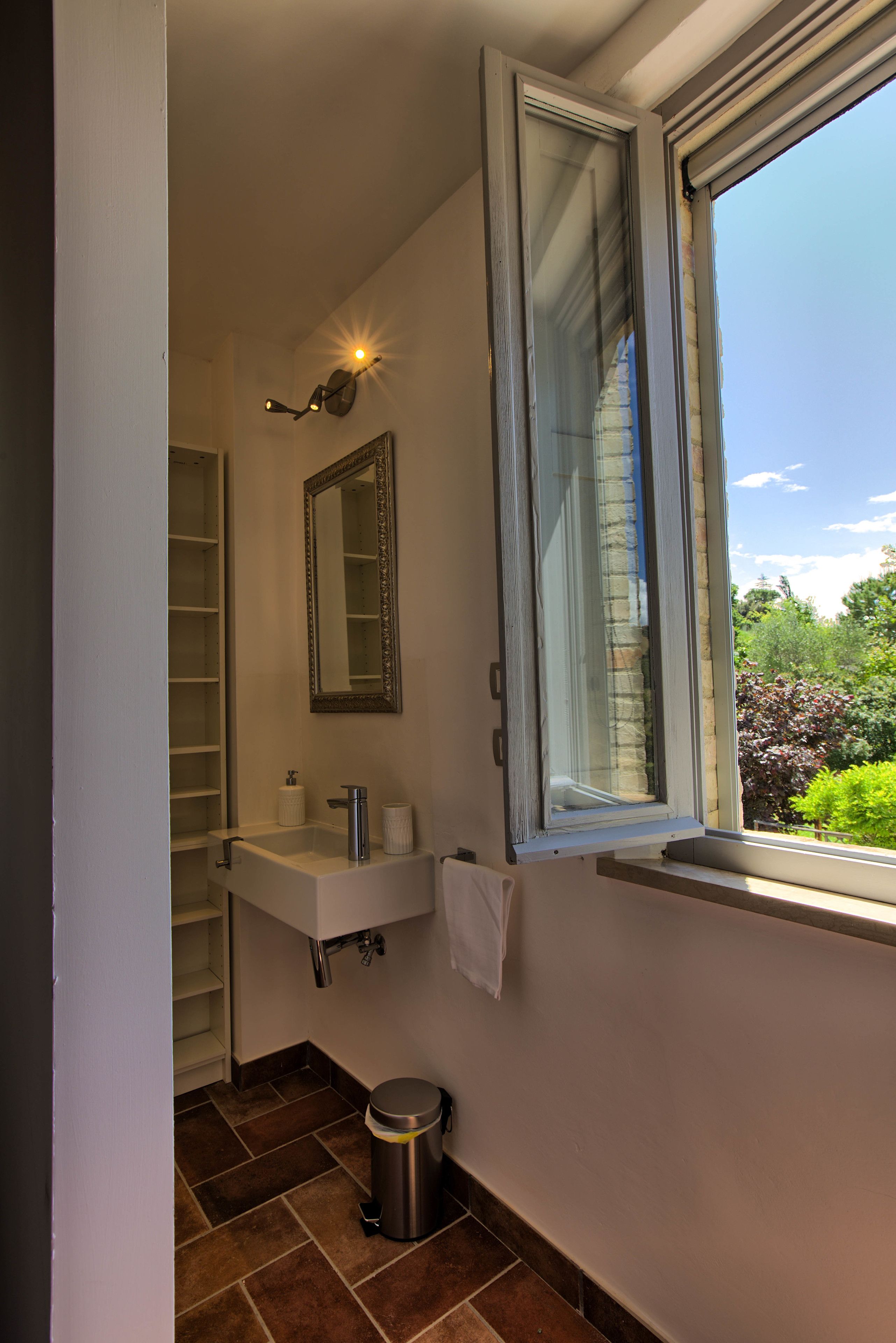 Un baño con un lavabo blanco, un espejo, una ventana y un cubo de basura.