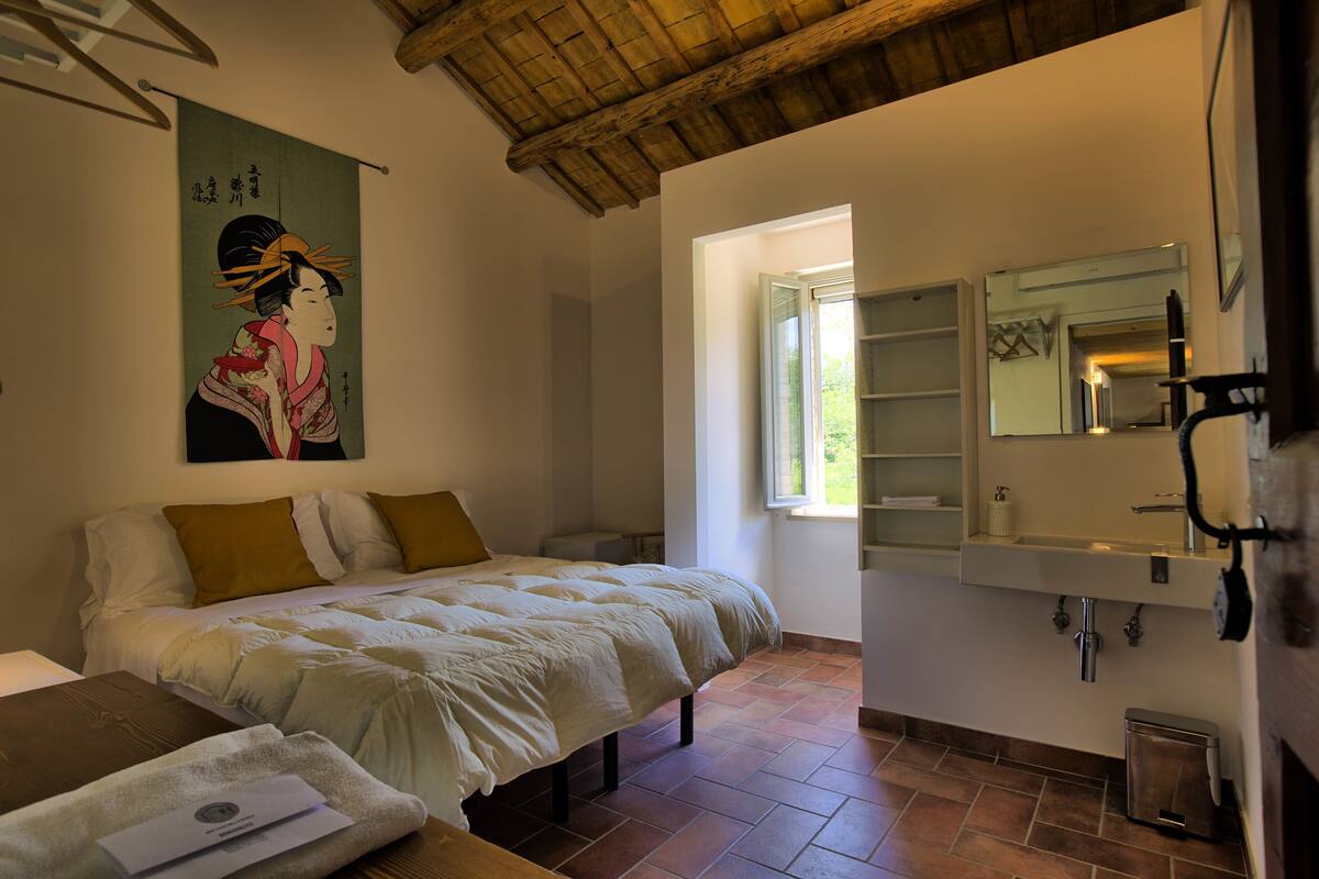 A bedroom with a bed, a wooden headboard, a large window, and a bathroom with a sink and mirror.