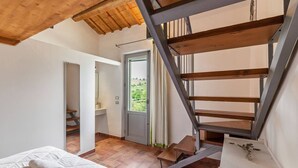 Une chambre avec un plafond en bois, un lit recouvert de linge de lit blanc, un escalier avec des marches en bois et une vue sur la verdure à l’extérieur.