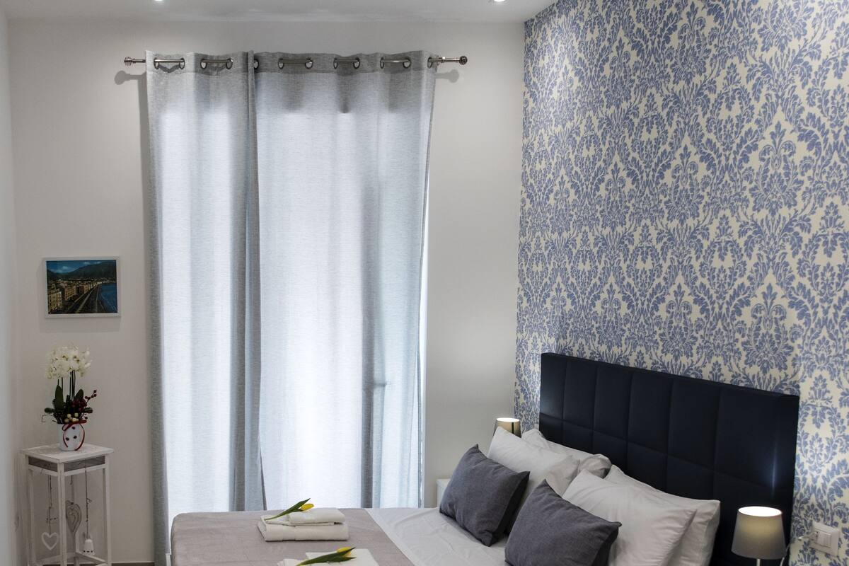 A modern bedroom with a large bed, a bedside table, a nightstand, and a wall with a floral pattern.