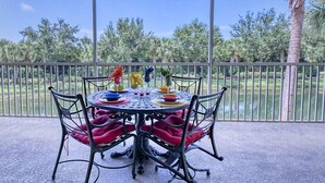 A covered patio with a table set for four, surrounded by metal chairs with red cushions, overlooking a landscaped area with trees and a palm tree. - Sabal Pointe at Majestic Palms 1004 Near beach Townhome. Sanibel/Captiva/Fort (Fort Myers)