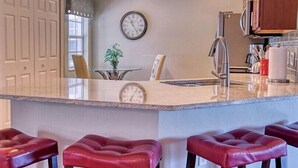 A kitchen with a marble island, red leather bar stools, a clock on the wall, and wooden cabinets. - Sabal Pointe at Majestic Palms 1004 Near beach Townhome. Sanibel/Captiva/Fort (Fort Myers)