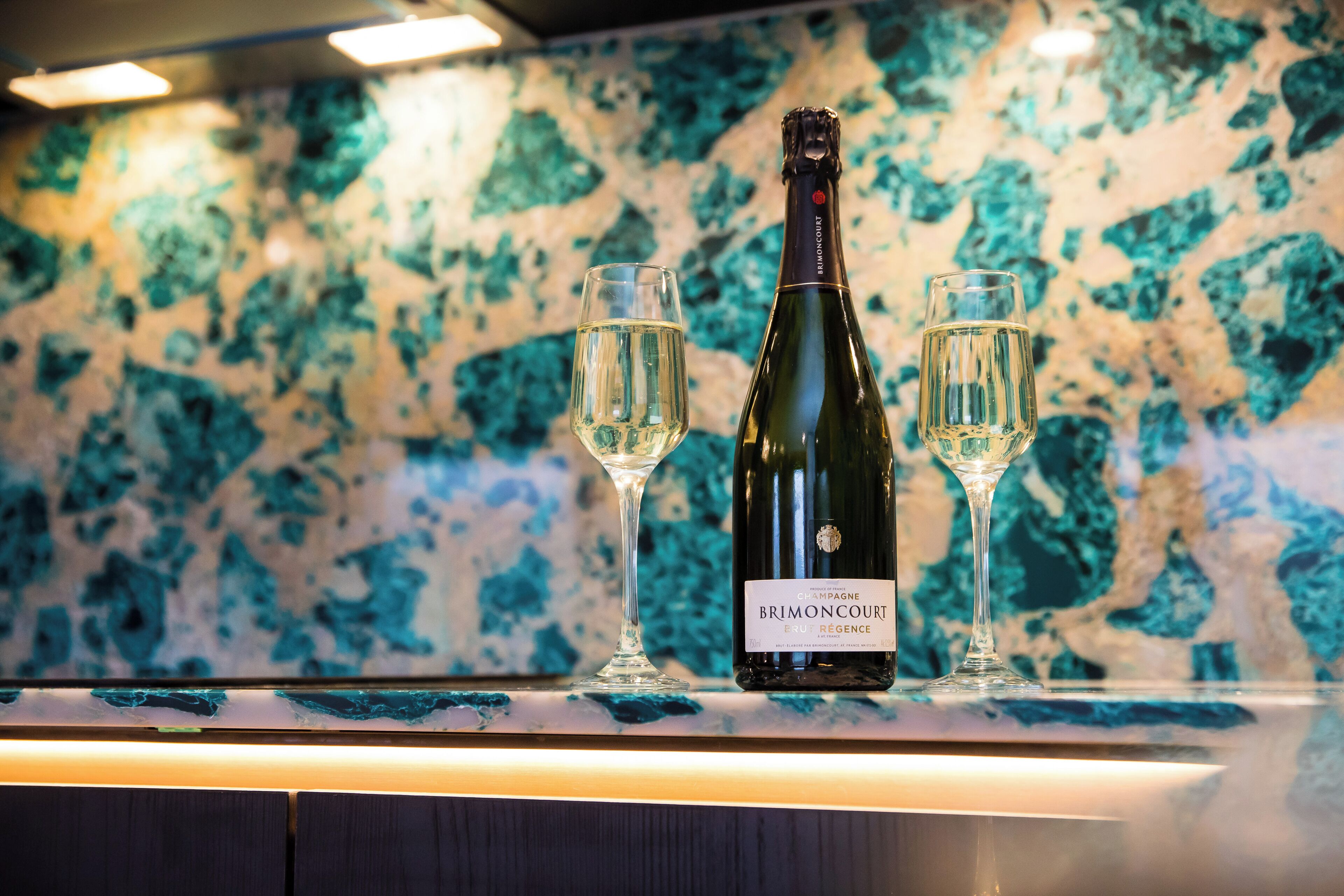 Eine Flasche Brimontcourt Champagner und zwei Gläser auf einer Theke mit Marmorrückwand.