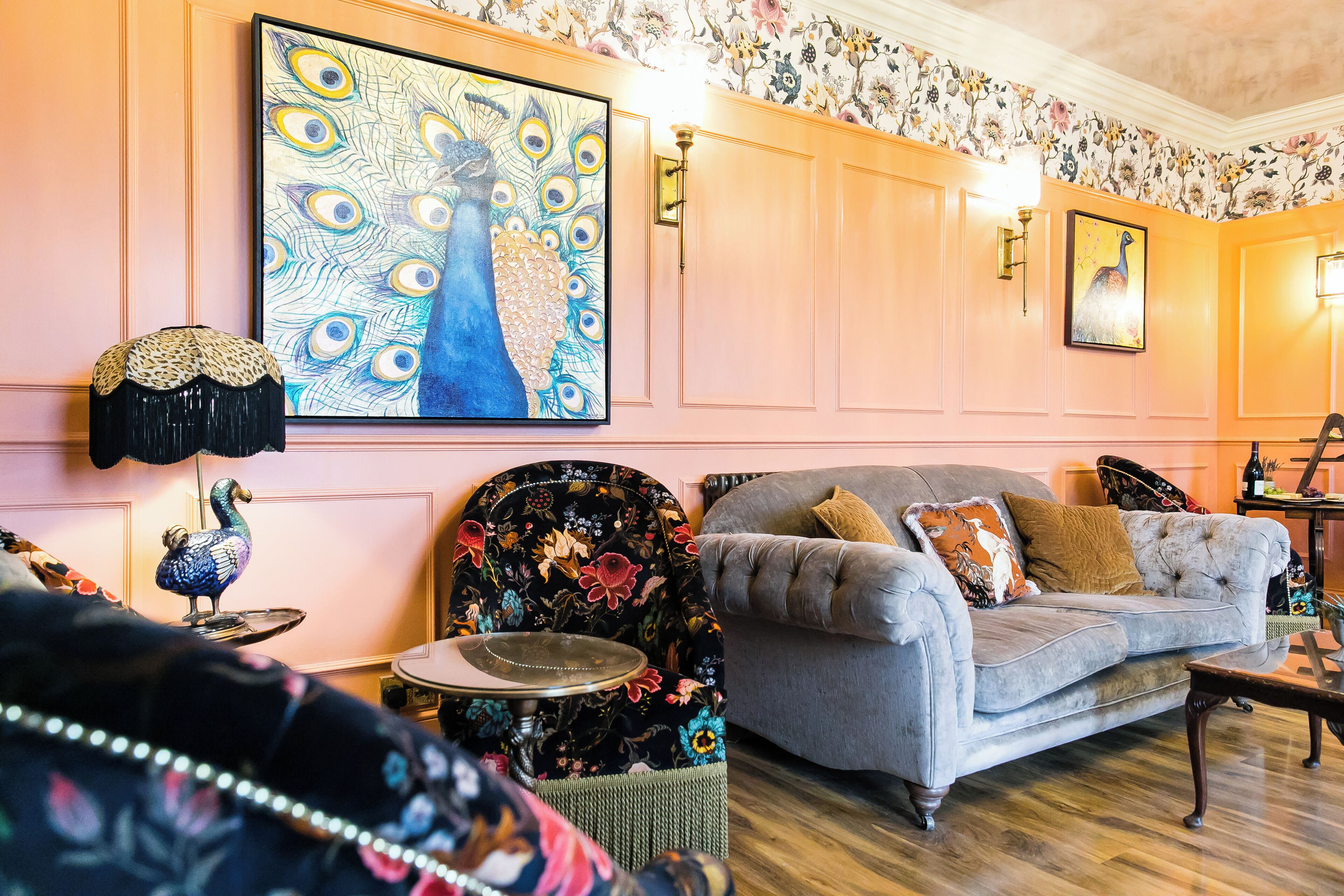 Een woonkamer met een pauwen schilderij, bloemen behang, een gestuufde bank en een decoratieve tafel.