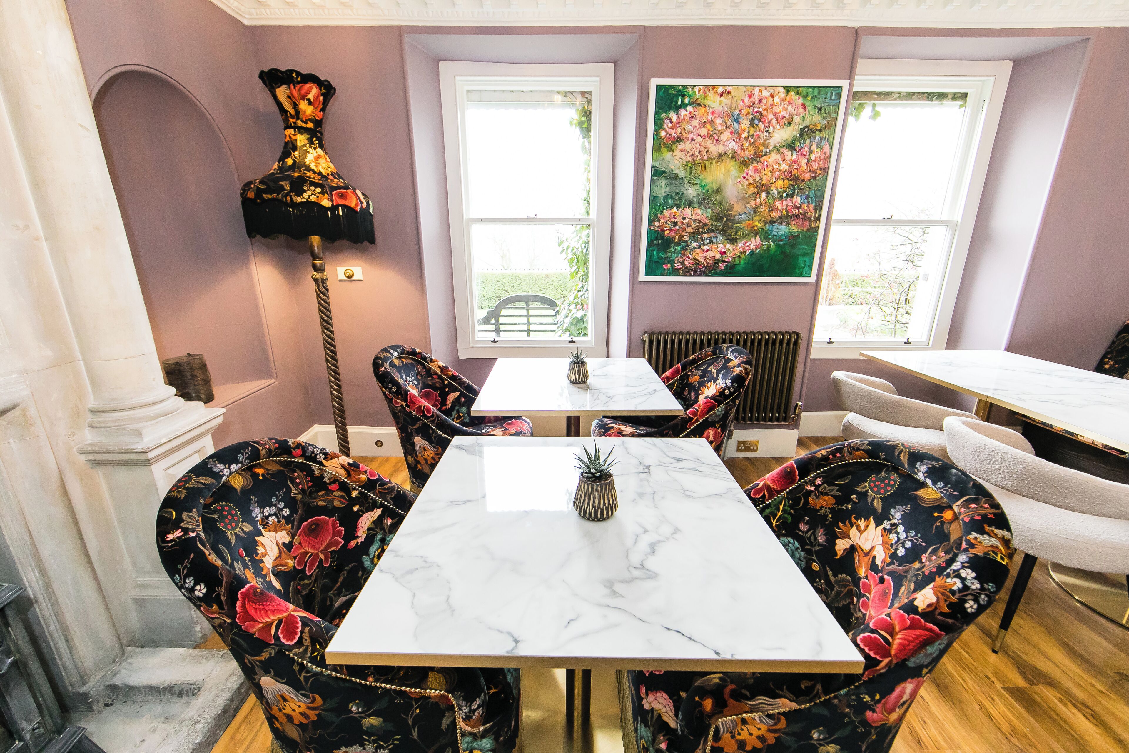 Un comedor con una mesa de mármol, sillas tapizadas con motivos florales, una lámpara de pie alta y un cuadro grande con flores en la pared.