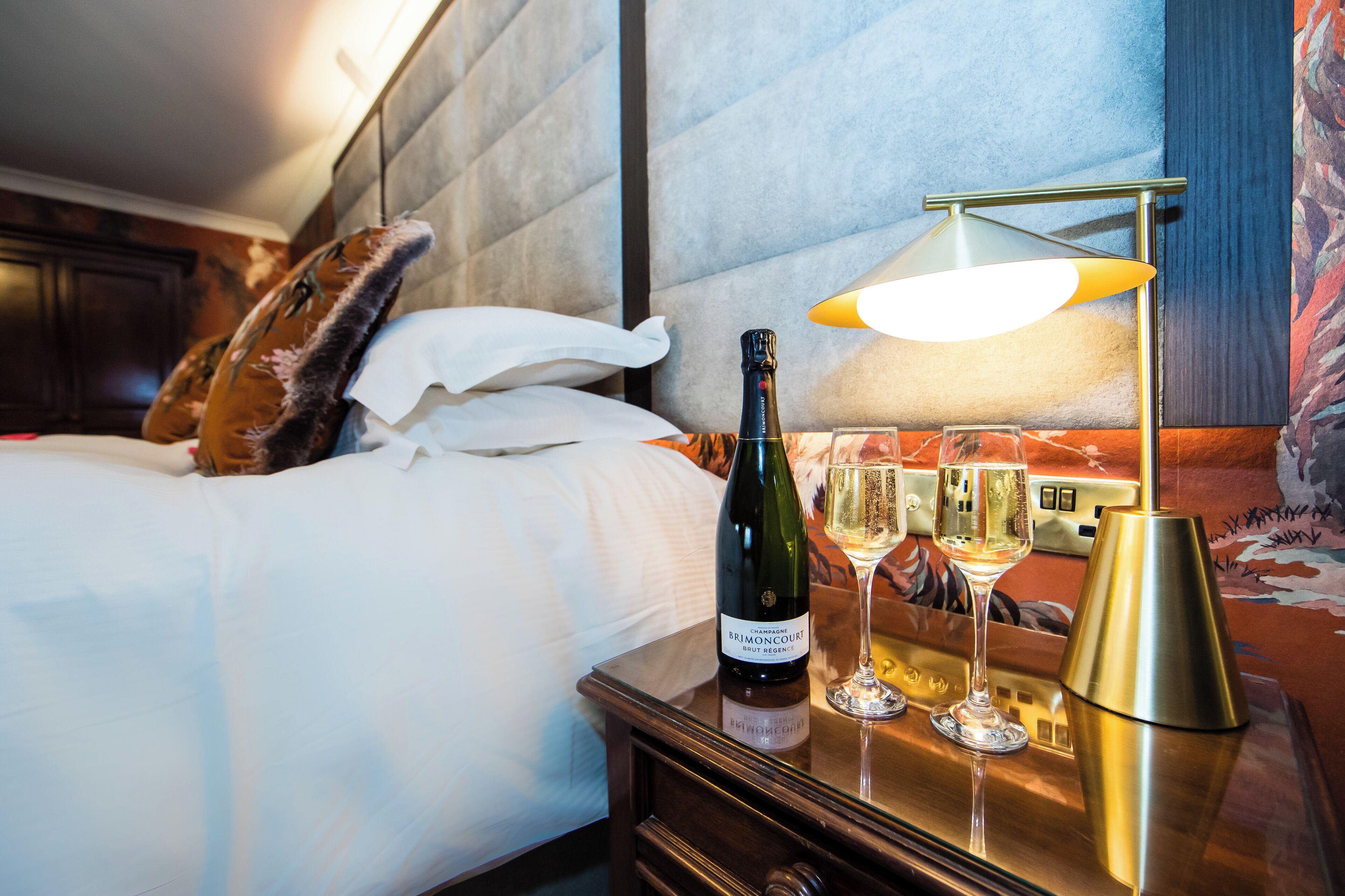 A hotel room with a bed, a bottle of wine, and two glasses on a nightstand.