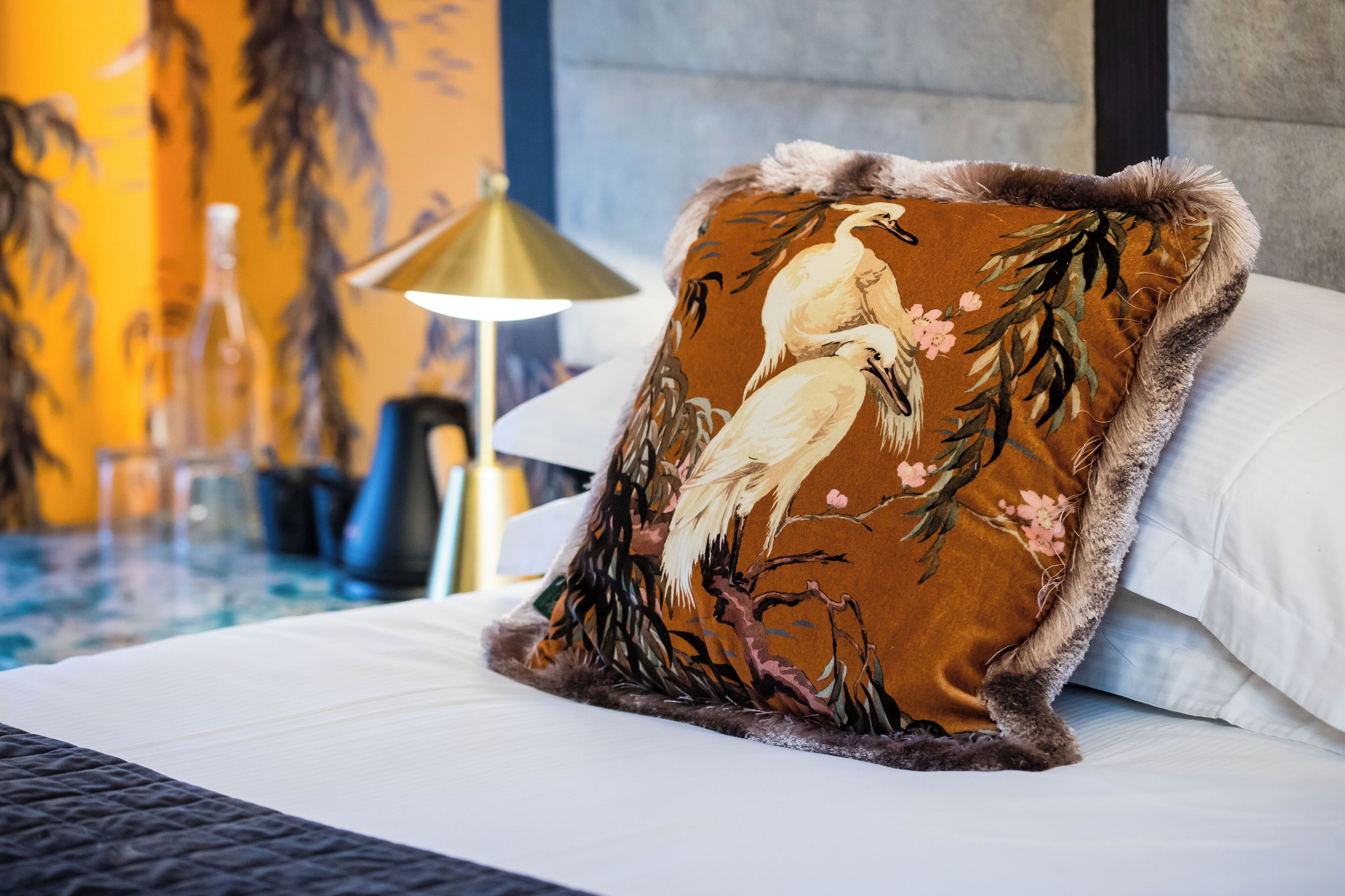 A bed with a decorative pillow featuring a bird and floral design, a lamp, and a wall with a mural.