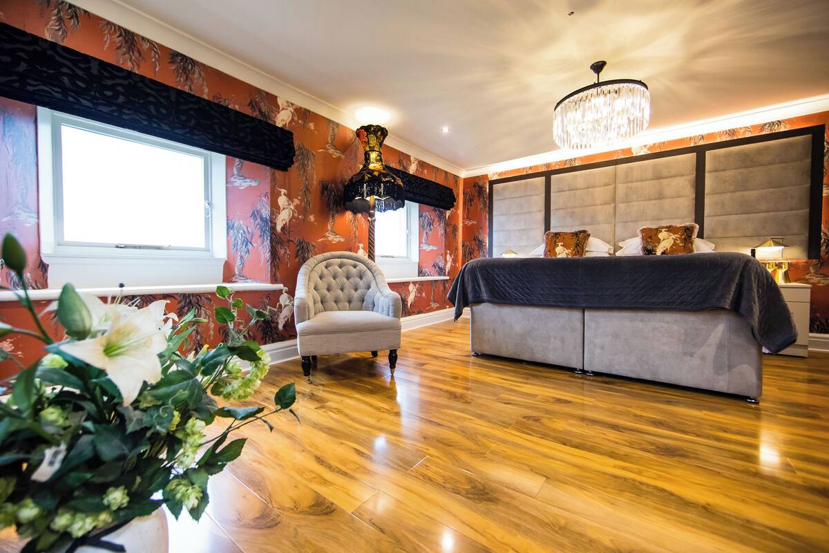 A bedroom with a large bed, a tufted armchair, a chandelier, and a window with curtains.