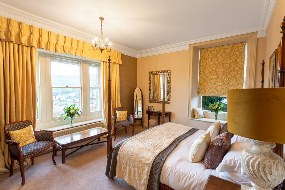 A hotel room with a large bed, a sitting area with a chair and a table, a window with curtains, a mirror, and a lamp.