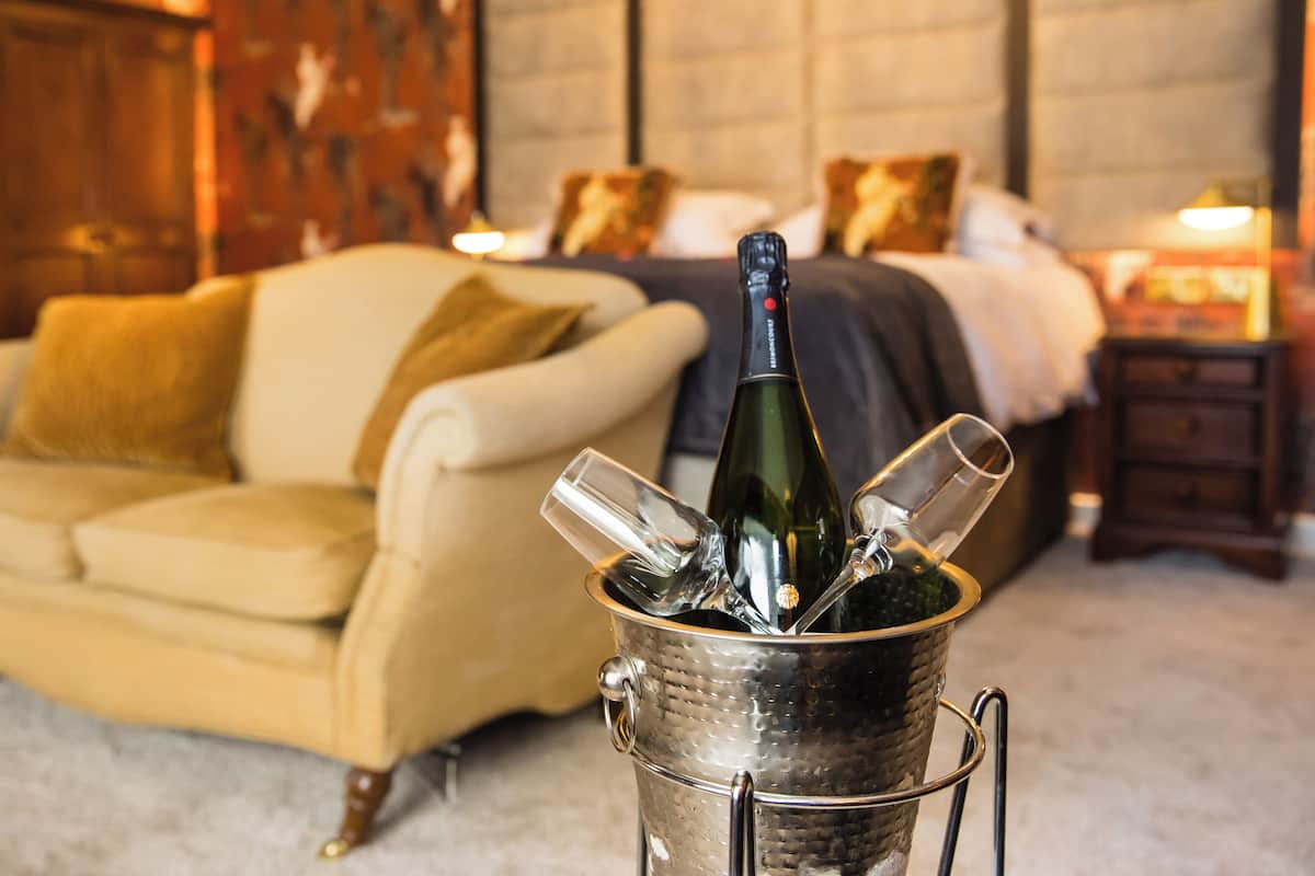 A room with a bed, a bedside table, a nightstand, a lamp, and a bottle of champagne in a metal bucket.