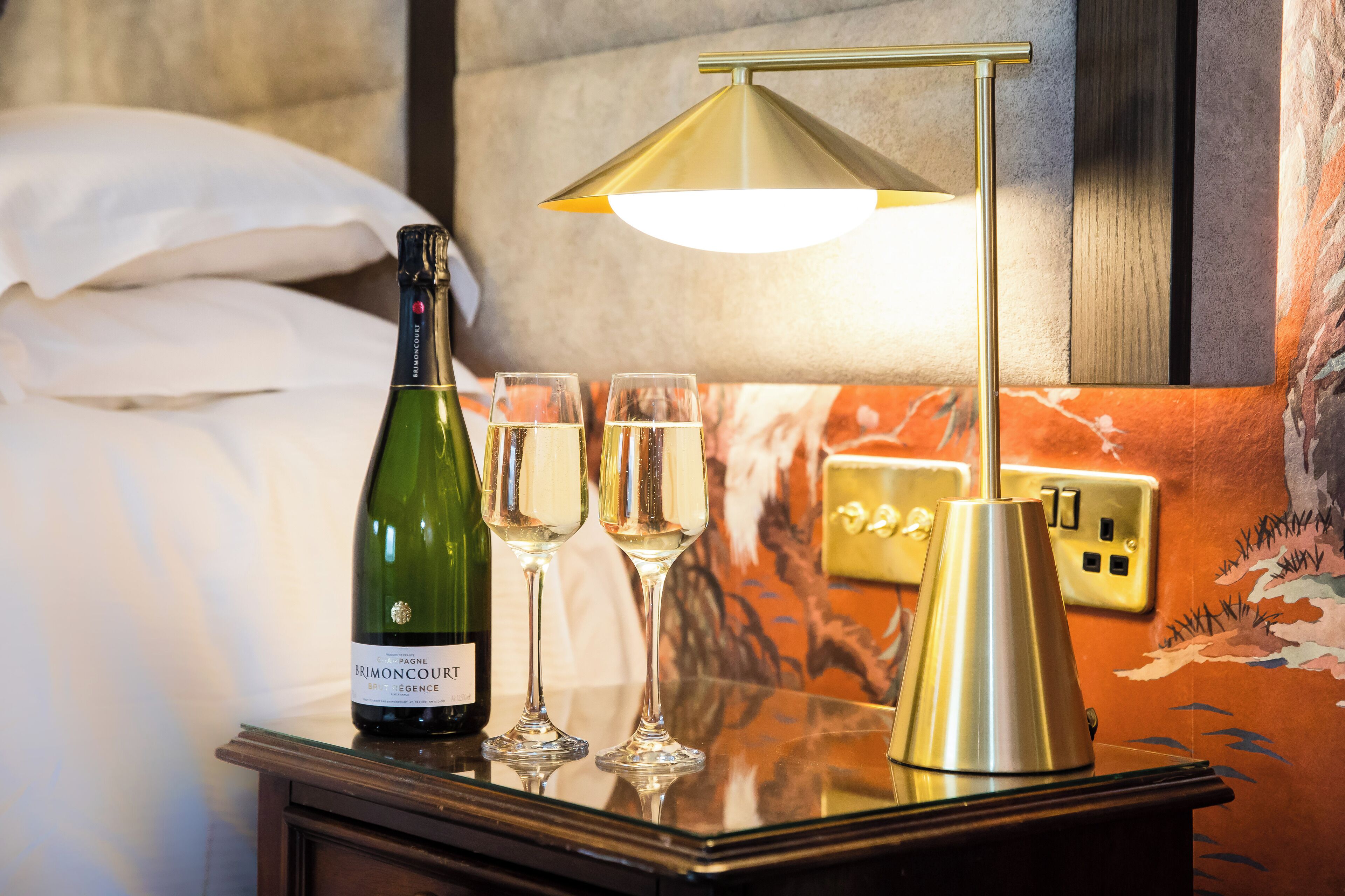 Una habitación de hotel con una cama, una mesita de noche con una botella de vino, dos copas y una lámpara.