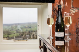 Una botella de champán Brut Regence Brimontcourt y dos copas sobre una mesa cerca de una ventana con vistas al paisaje.