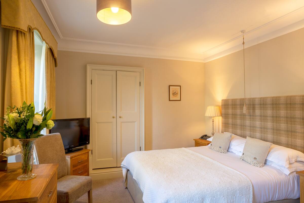 A hotel room with a bed, a desk with a chair, a television, a bedside table with a lamp, and a wardrobe.