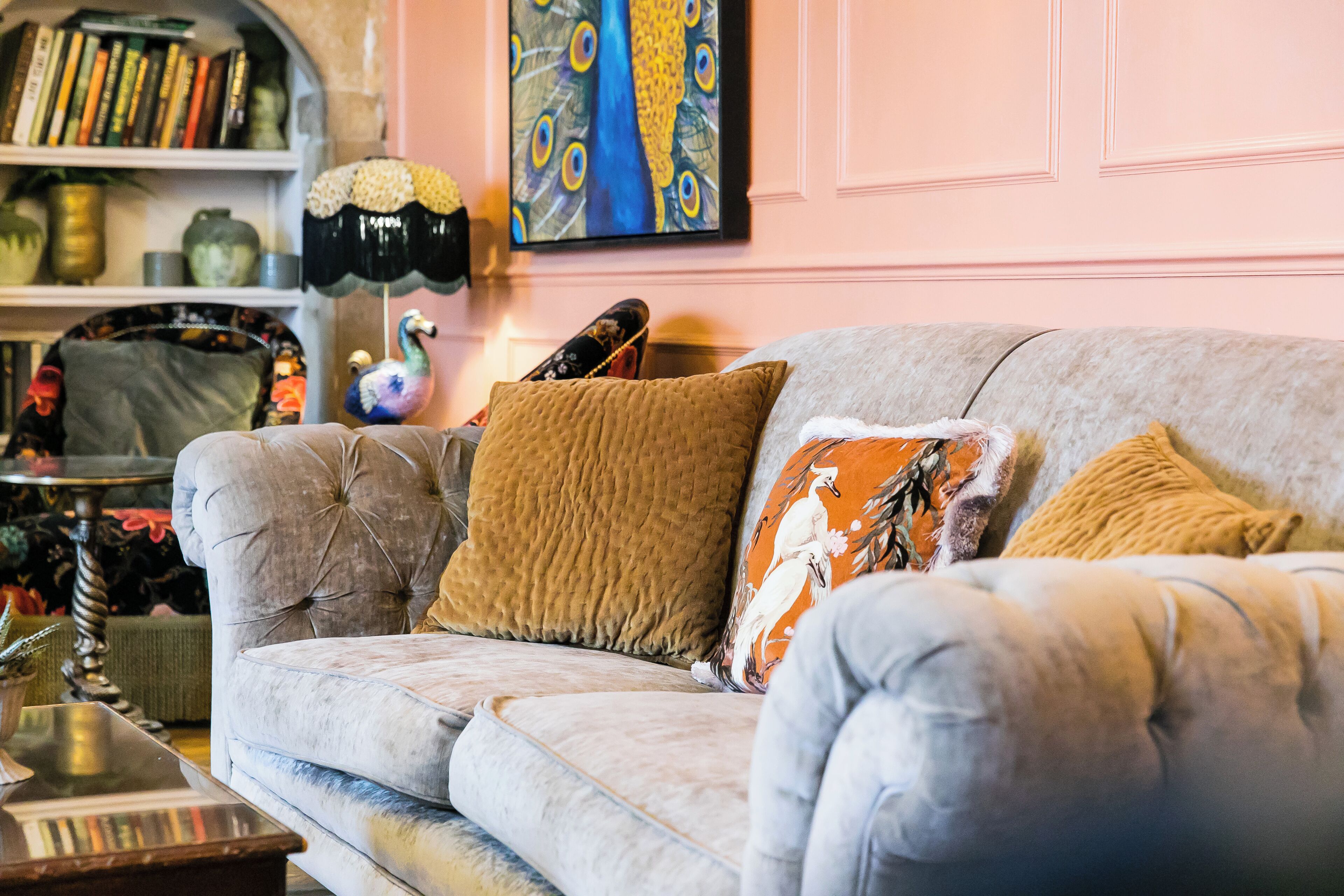 Un sofá mullido con varios cojines, un cuadro decorativo de un pavo real y una estantería con libros y objetos decorativos.