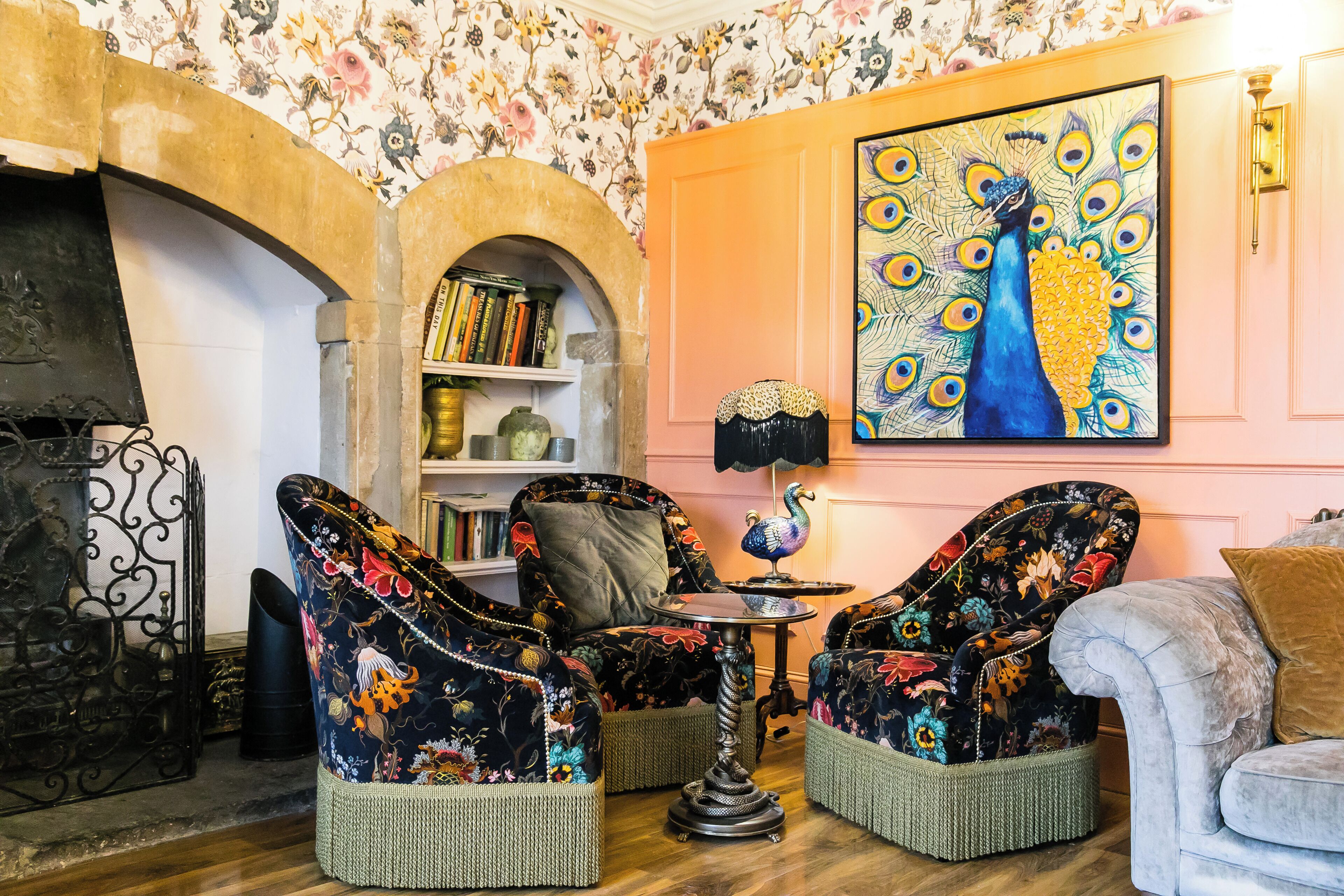 A cozy room with a fireplace, floral wallpaper, a peacock painting, and two upholstered chairs with a woven base.