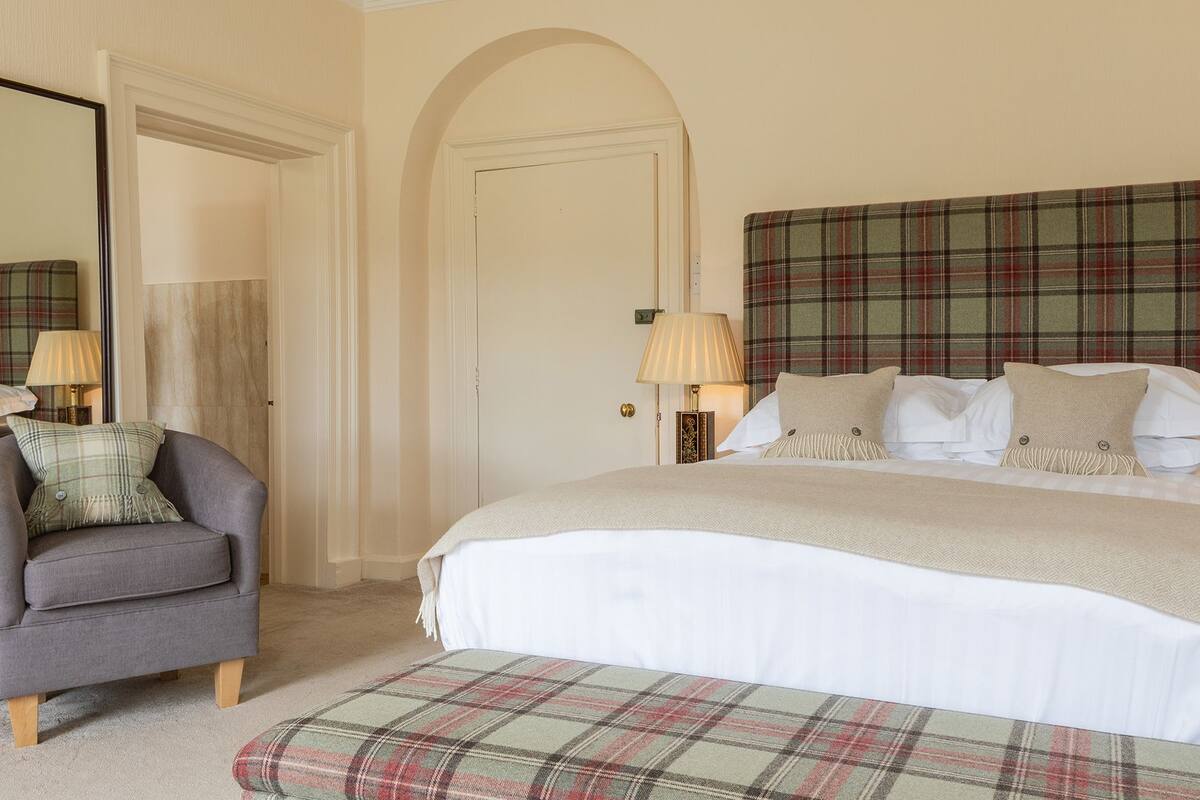 A bedroom with a large bed, a chair, a plaid headboard, and a mirror on the wall.
