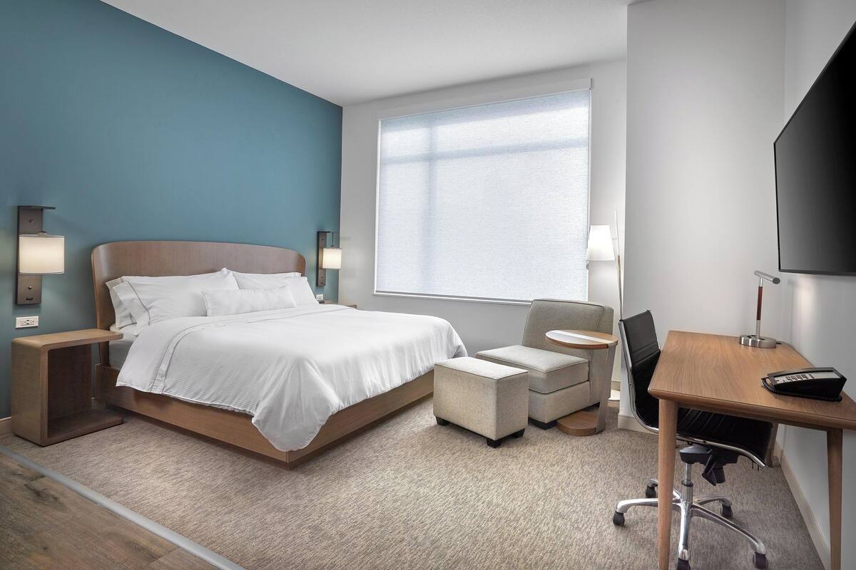A hotel room with a bed, a desk, a chair, a TV, and a window.