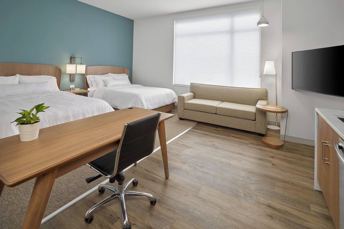 A hotel room with two beds, a desk with a chair, a sofa, a TV, and a window with blinds.