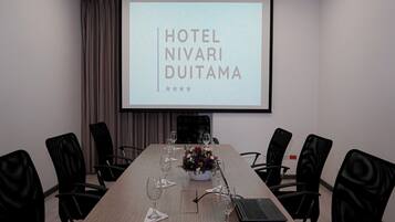 Un salón de conferencias con una larga mesa, sillas negras, una computadora portátil y una pantalla que muestra "HOTEL NIVARI DUTAMA".