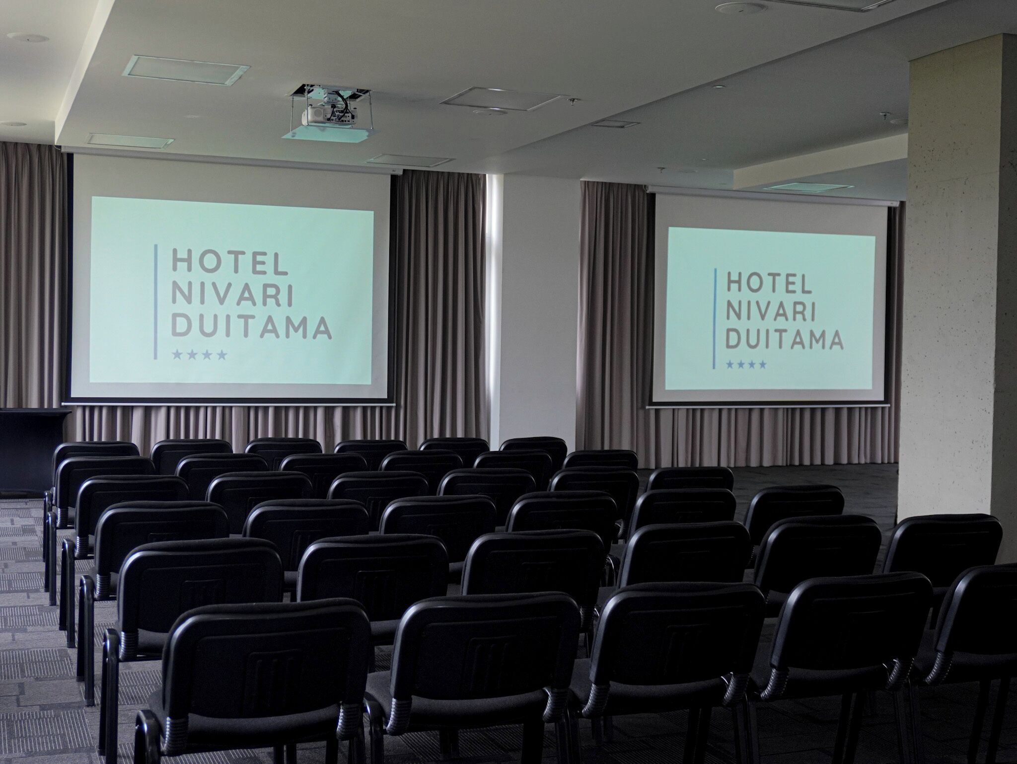 Ein Konferenzraum mit Reihen schwarzer Stühle und zwei Bildschirmen, auf denen 'HOTEL NIVARI DUITAMA' angezeigt wird.