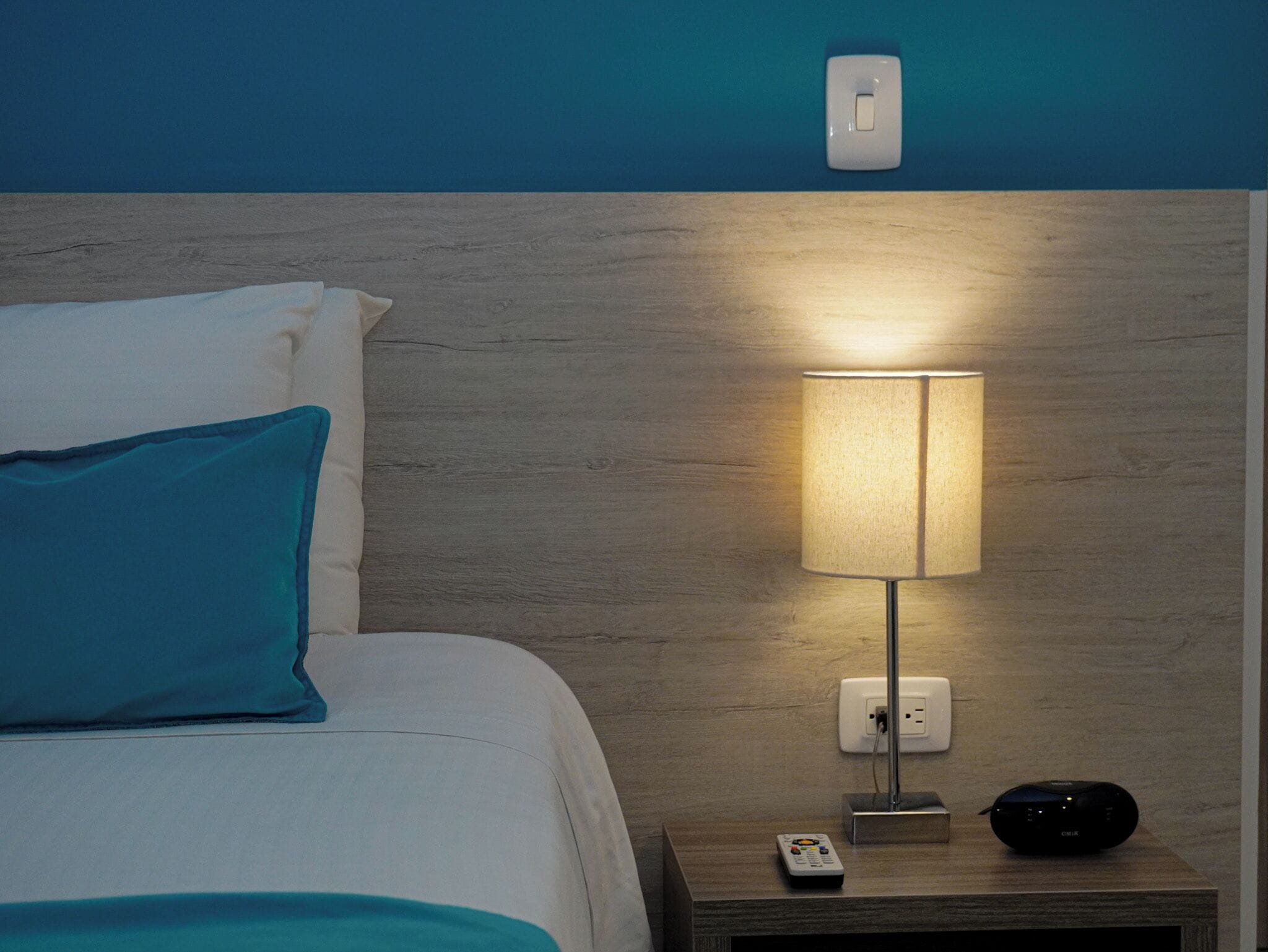 A neatly made bed with a teal pillow, a bedside lamp, a remote control, and a speaker on a wooden nightstand.