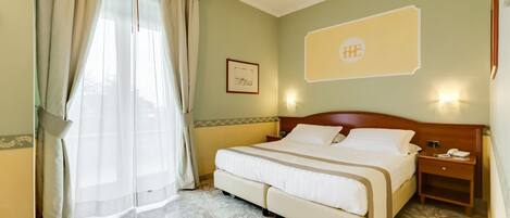A hotel room with a large bed, a nightstand with a phone, a framed picture on the wall, and a window with curtains.