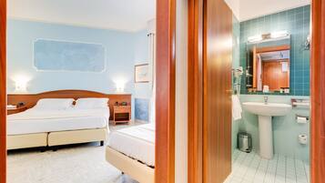 A hotel room with a double bed, bedside tables, a nightstand, a wall-mounted light, and a framed picture. The bathroom has a sink, mirror, towel rack, and a trash bin.