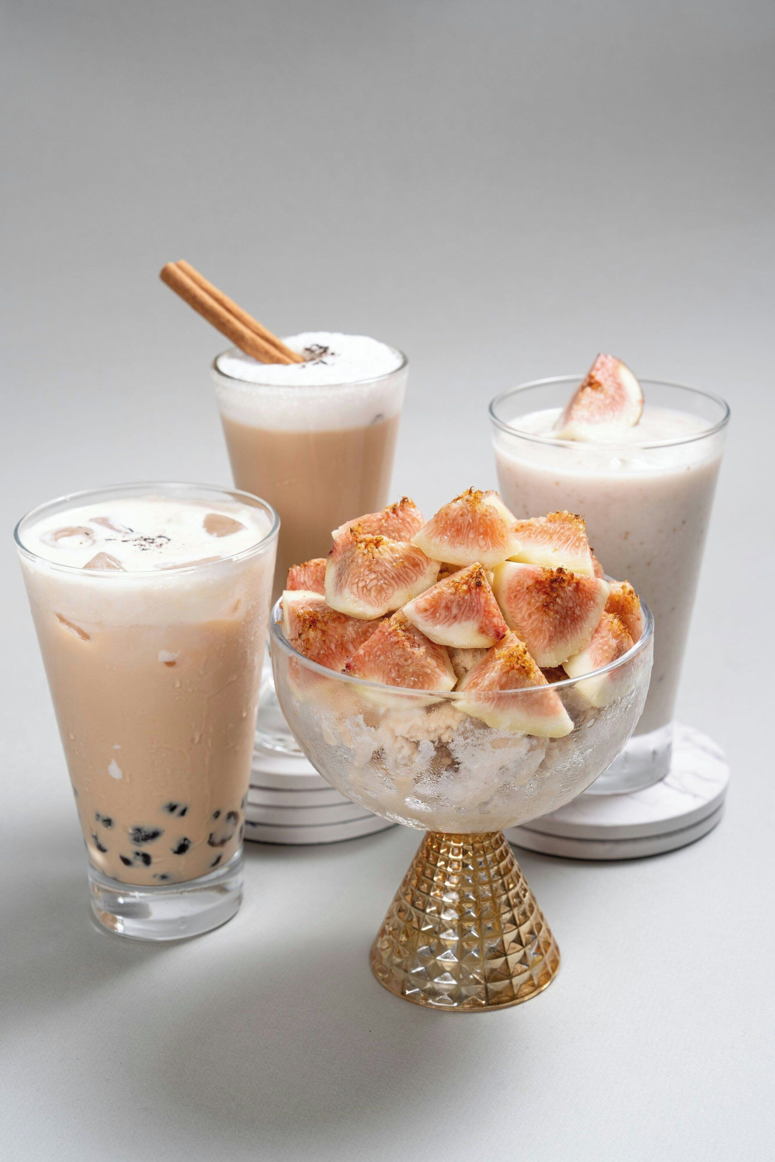 Three glasses of a creamy beverage, one with a cinnamon stick, and a bowl of fruit pieces on a decorative stand.