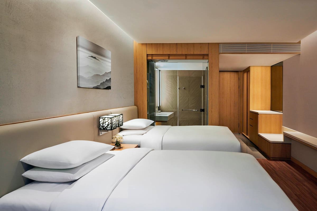 Ein Hotelzimmer mit zwei Betten, einem Holzschrank, einem Badezimmer mit Glastür und einem an der Wand hängenden Kunstwerk.