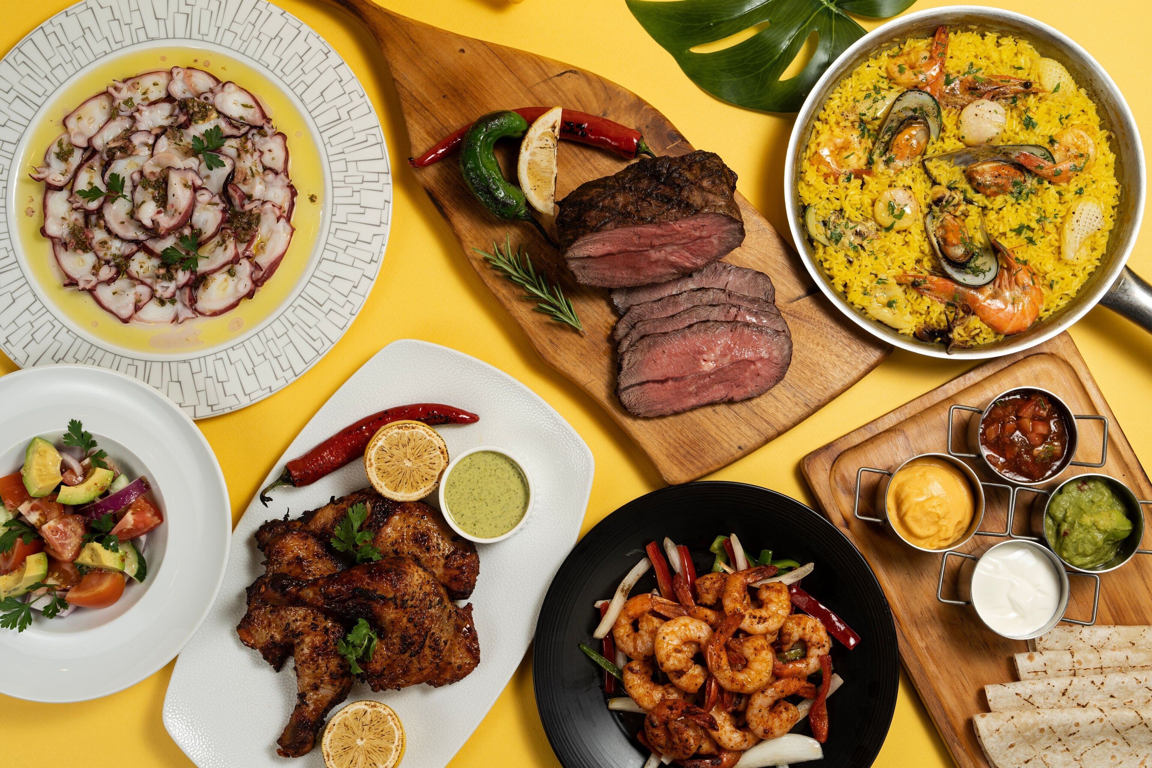 A variety of dishes including seafood, grilled meat, and rice dishes, served on multiple plates and a wooden board.
