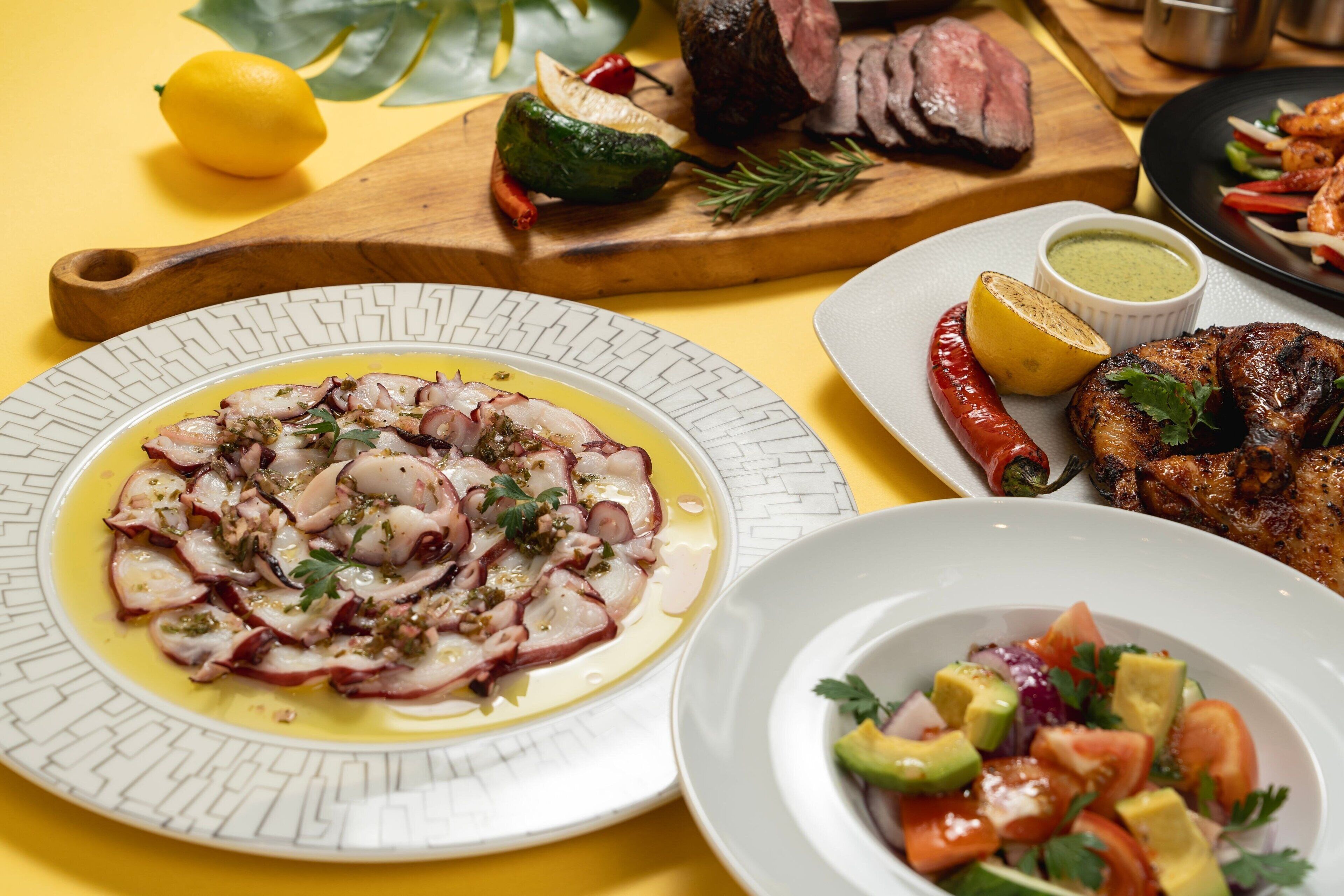 A table with various dishes including grilled meat, octopus salad, and a vegetable salad.