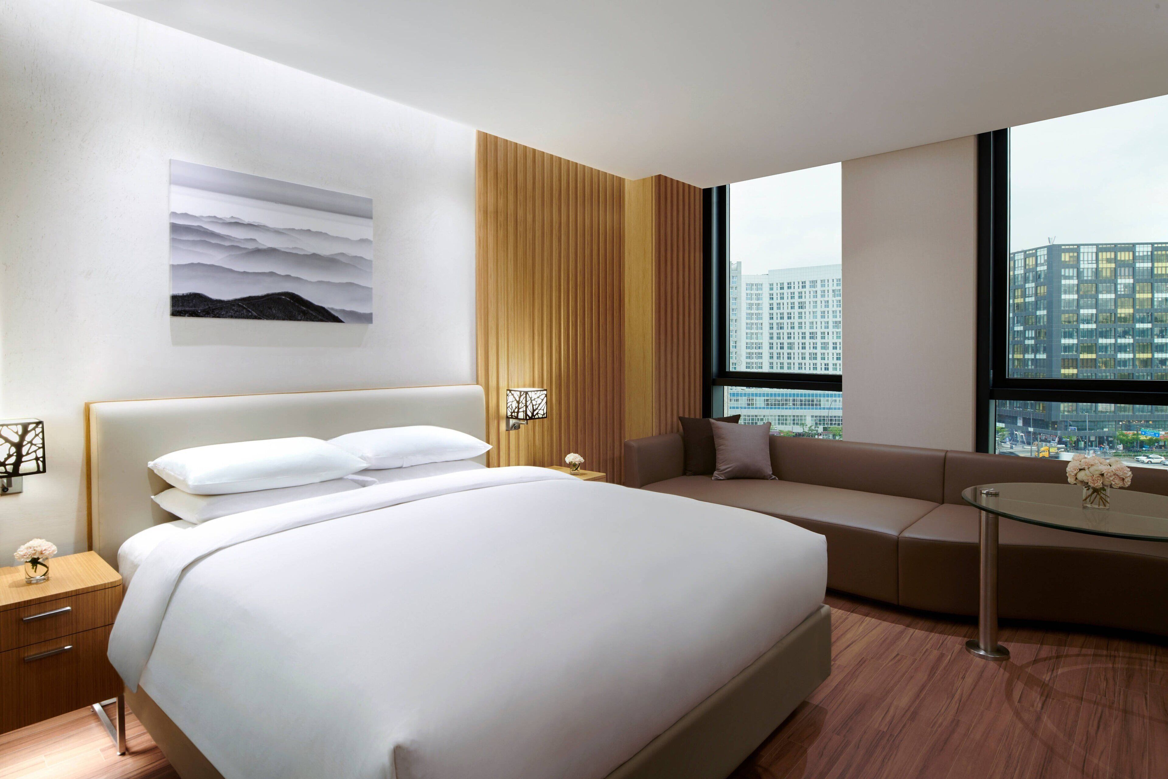 A modern hotel room with a large bed, a sofa, a small table, and a city view through large windows.