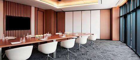 A conference room with a long table, white chairs, and a flat-screen TV.