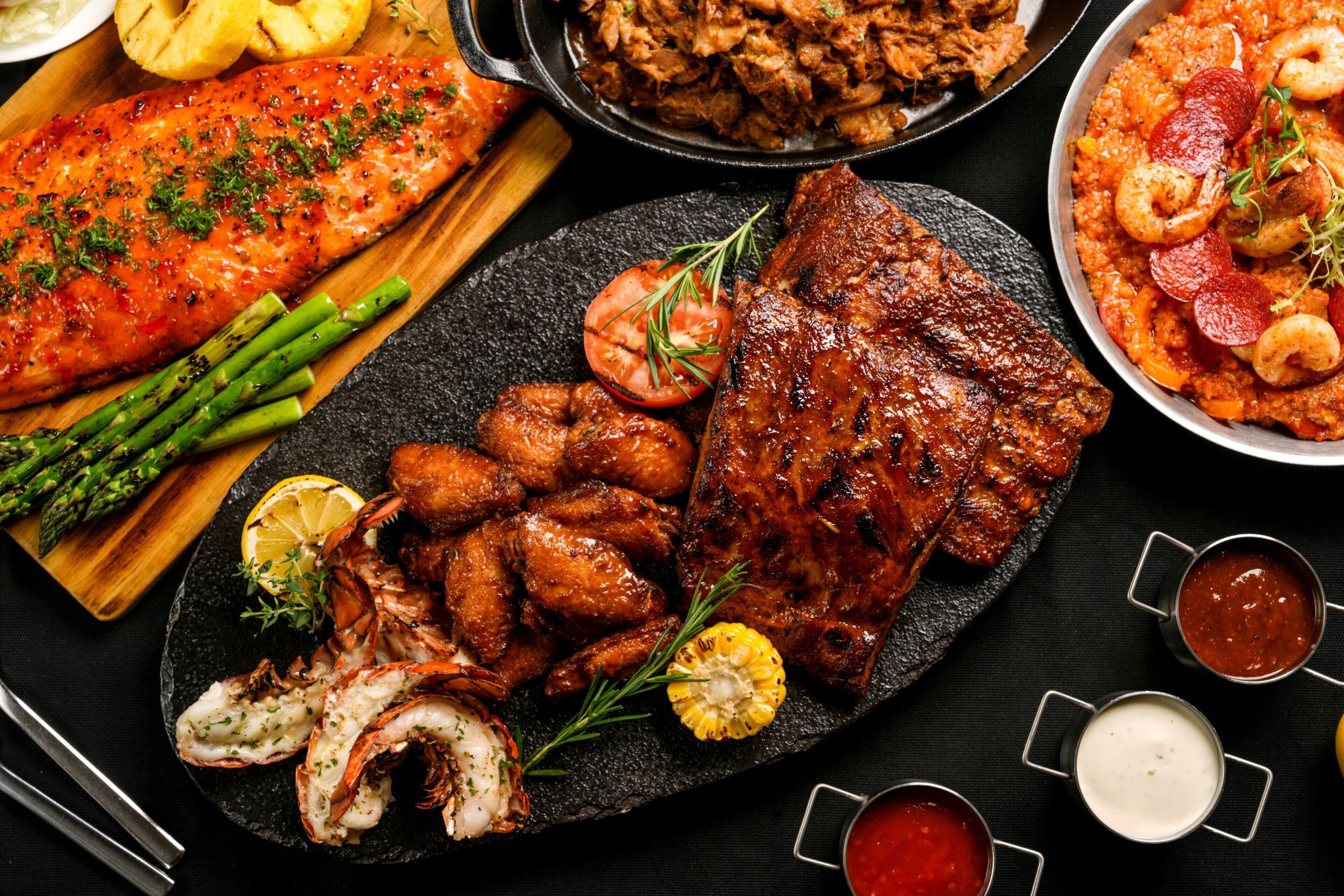 A variety of grilled meats including salmon, chicken wings, and ribs, served with vegetables and garnishes.
