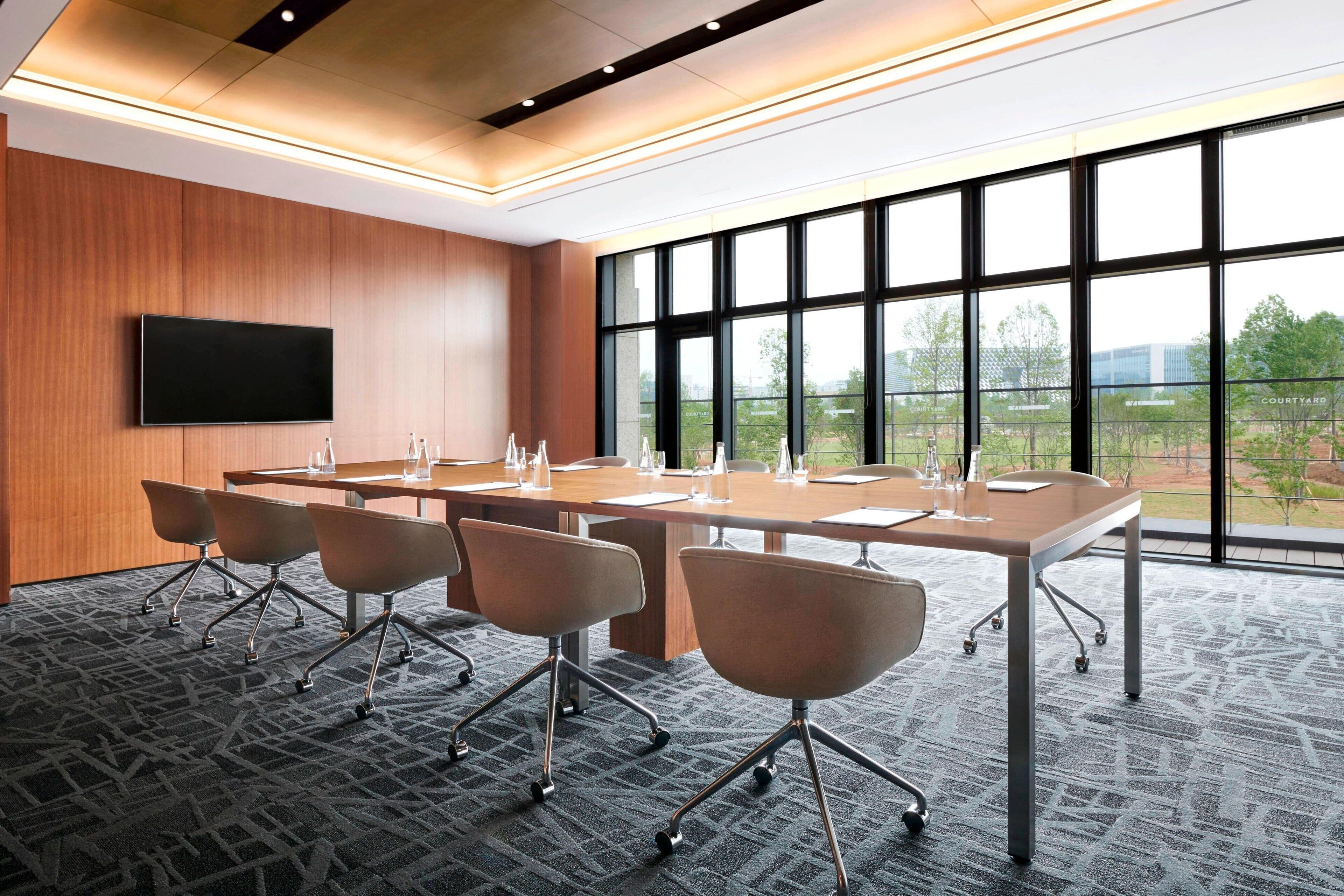 A conference room with a long table, four chairs, a flat-screen TV, and large windows offering a view of the outdoors.