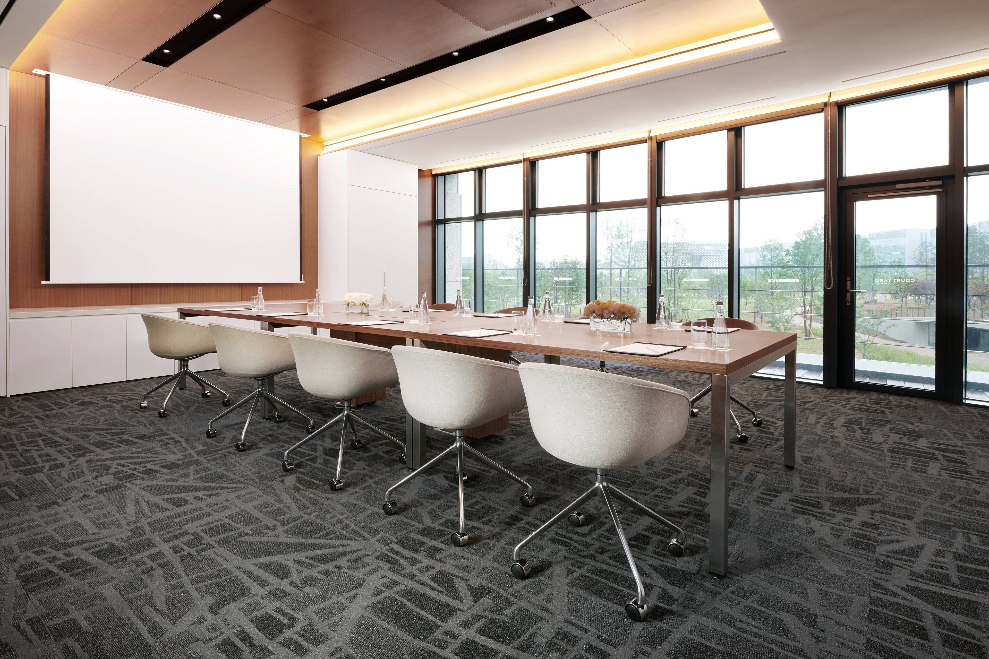 A conference room with a large table, four chairs, a projector screen, and large windows offering a view of the outdoors.