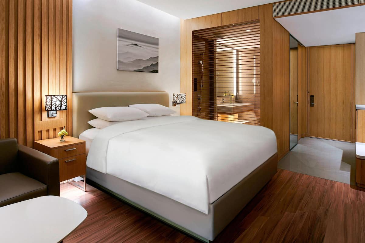 Ein modernes Hotelzimmer mit einem großen Bett, Holzvertäfelung, einem Nachttisch mit Lampe und Blick durch eine Schiebetür.