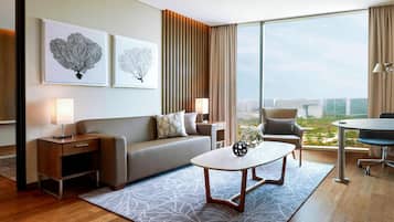 A modern hotel room with a large window, a sofa, a coffee table, and a desk with a lamp.