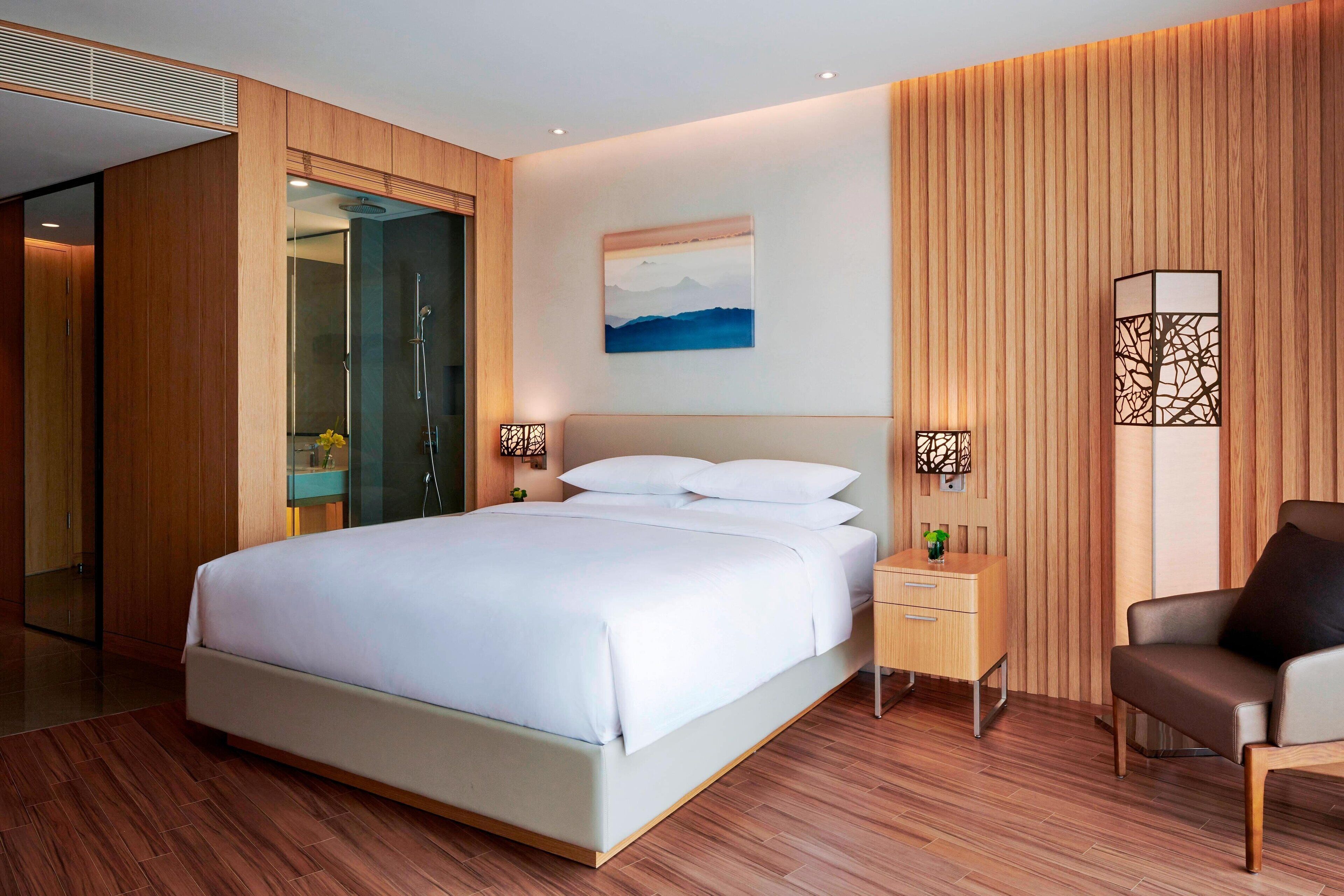 A modern hotel room with a large bed, wooden panel walls, a painting, and a nightstand with a lamp.