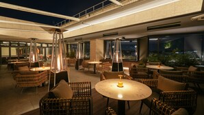 Een terras met rieten meubels en tafels, met uitzicht op de stad in de avond.