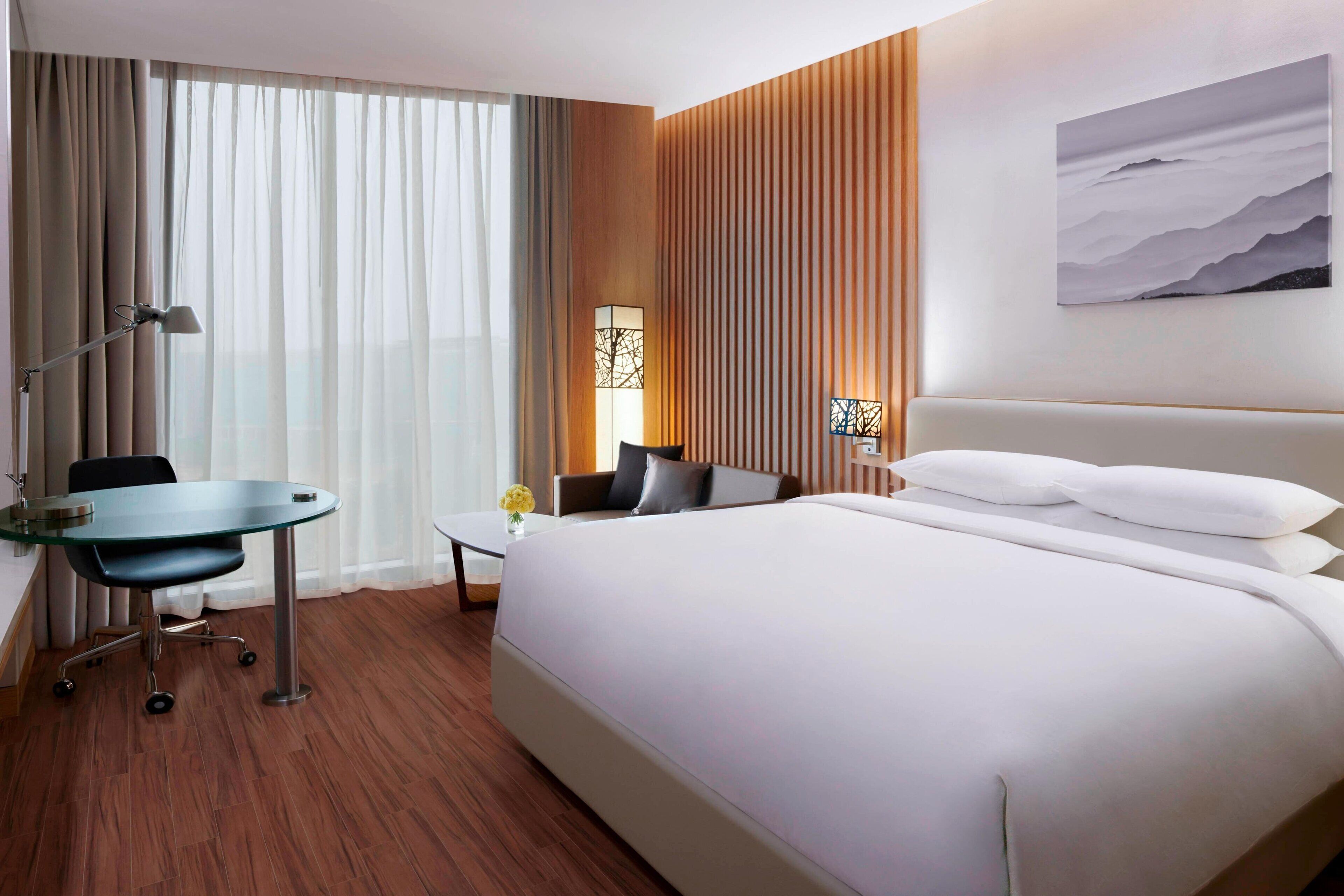 Habitación de hotel con una cama grande, un escritorio con una silla, una mesita con un florero y una lámpara de pared.