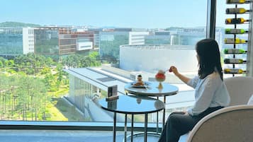 Una mujer sentada junto a una ventana con vista a la ciudad, una mesa con un postre y una estantería con vinos.