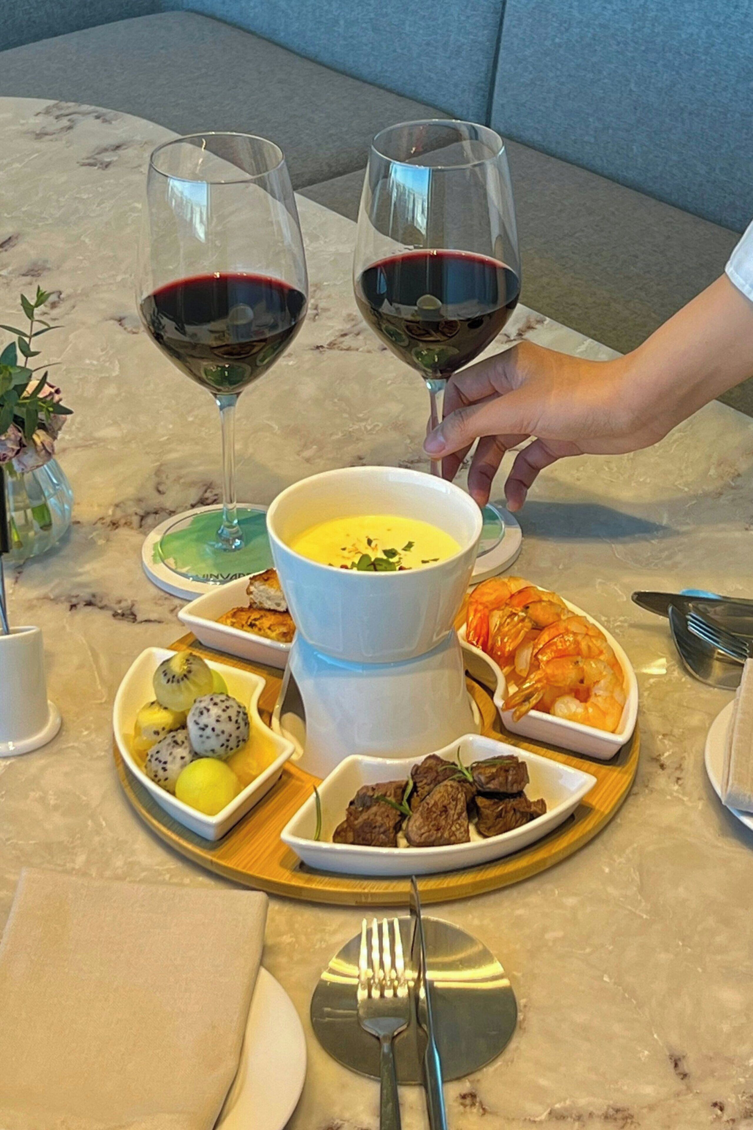 Une table à manger dressée avec une variété de plats, notamment des crevettes, de la viande et des fruits, ainsi que deux verres de vin rouge.
